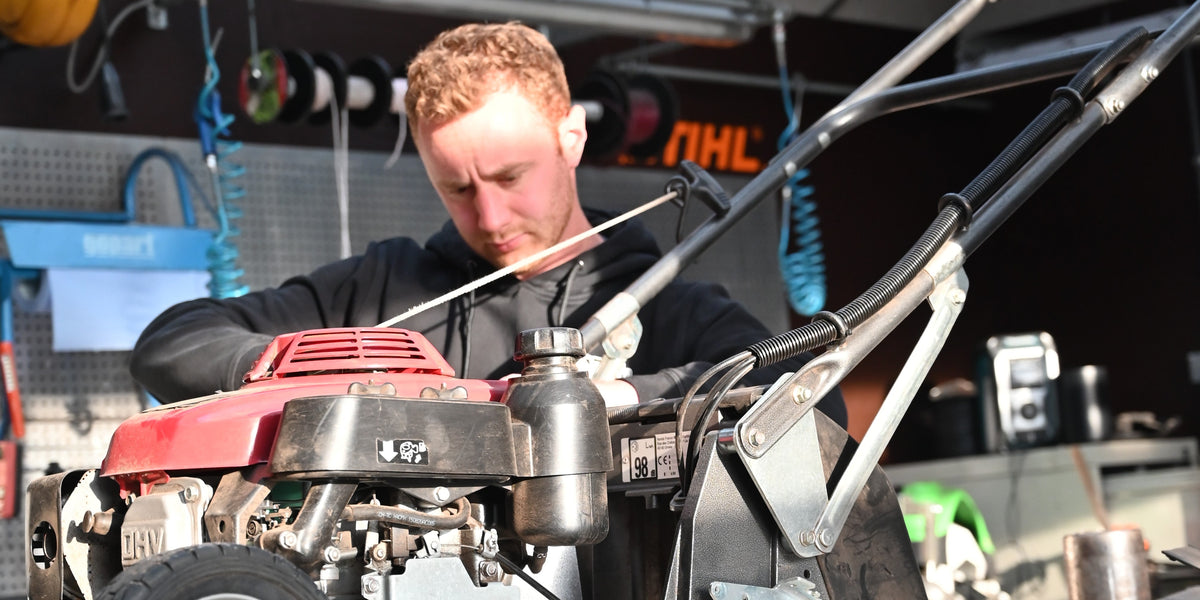 A man in a black hoodie works on a red and black push lawn mower inside a workshop or garage setting.