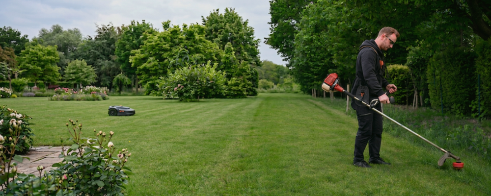 A person trims the lawn edge with a grass trimmer while, in the background, a robotic mower like one from Segway or Honda is moving across the lawn.