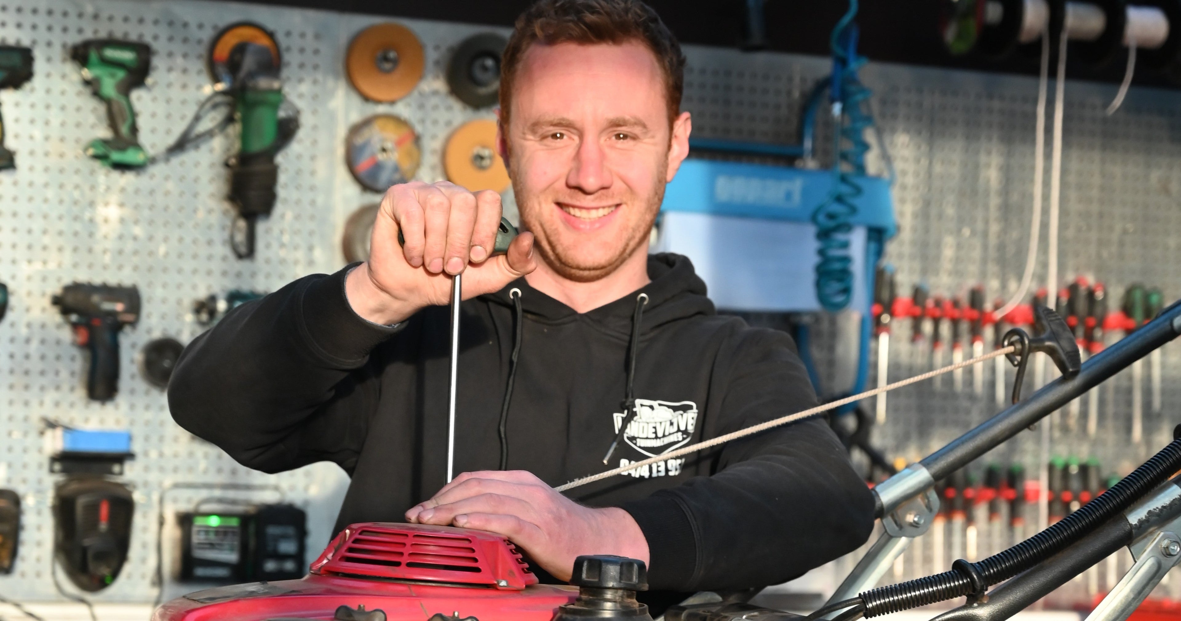 A man in a black hoodie stands behind a red and black lawnmower, holding the dipstick handle. The background shows a workshop pegboard with various tools.