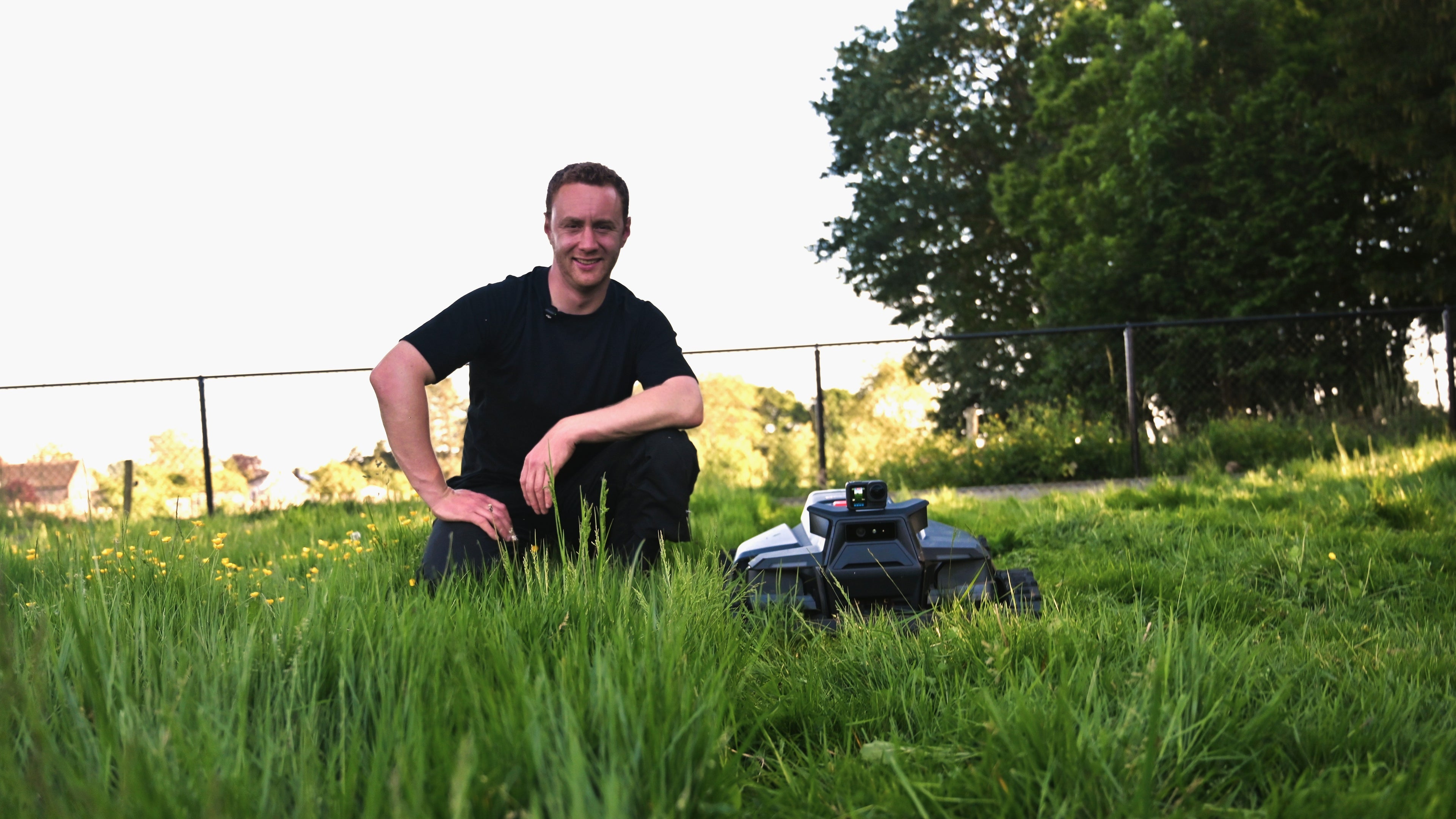 Person kneeling in grass next to a lawn mower with trees and fence in the background